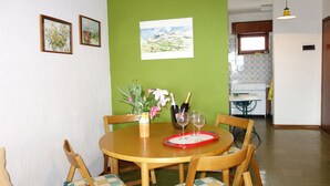 Un comedor con una mesa redonda de madera, cuatro sillas y un florero con flores. Hay cuadros en la pared y una ventana al fondo.