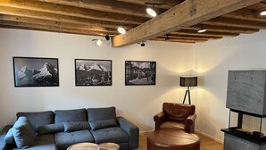 Ein modernes Wohnzimmer mit einem grauen Ecksofa, einem braunen Lederhocker, zwei Holztischen und einer Stehlampe. An der Wand befinden sich drei gerahmte Fotografien und ein Fernseher auf einem Schrank.