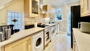 A modern kitchen with a built-in washer and dryer, wooden cabinets, and a large window with a view of greenery. - Stylish 3-bed House in Hornchurch - Free Parking (Hornchurch)