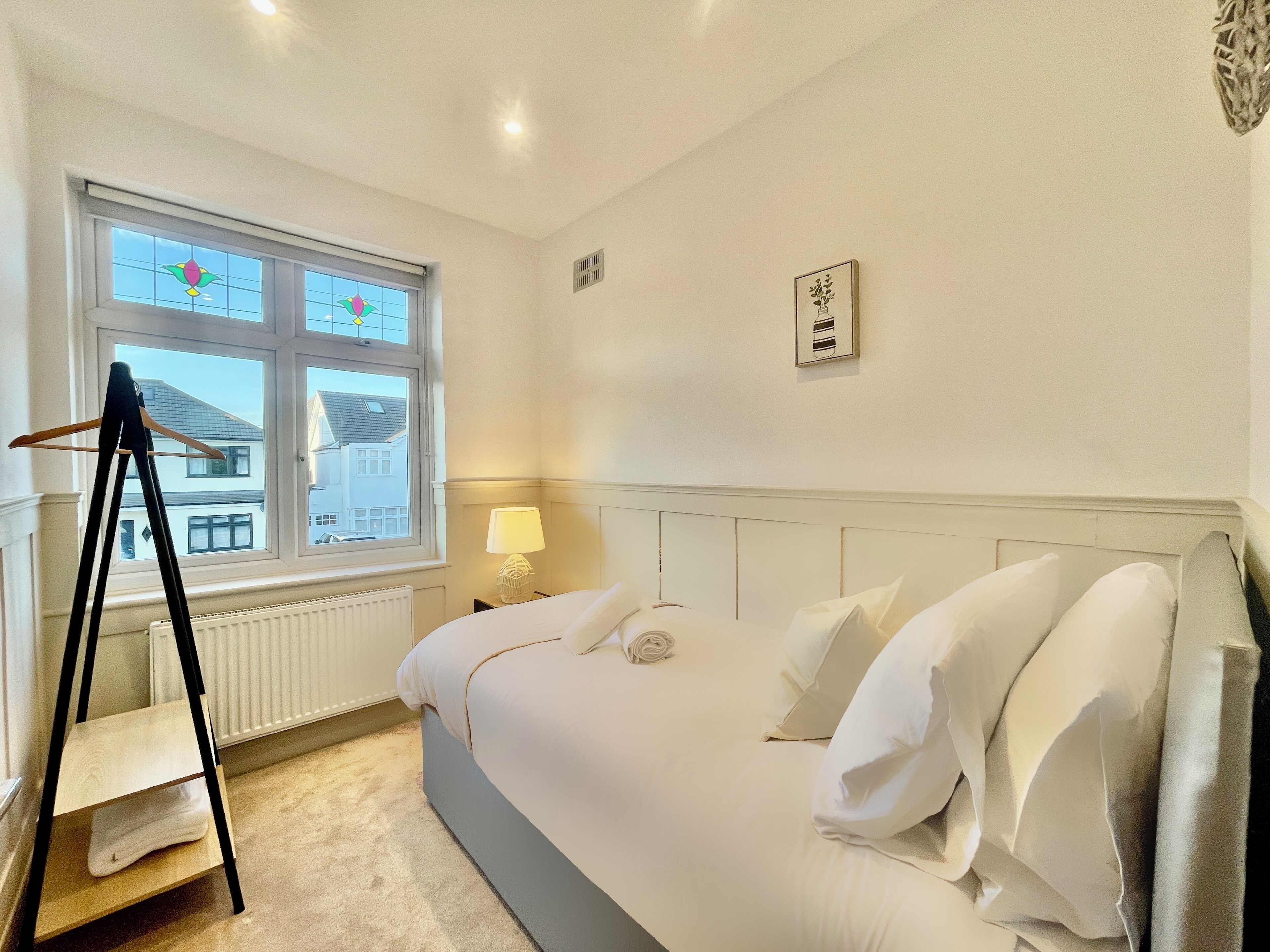 A bedroom with a bed, a window, a radiator, a wall-mounted air conditioning unit, and a bedside table with a lamp.