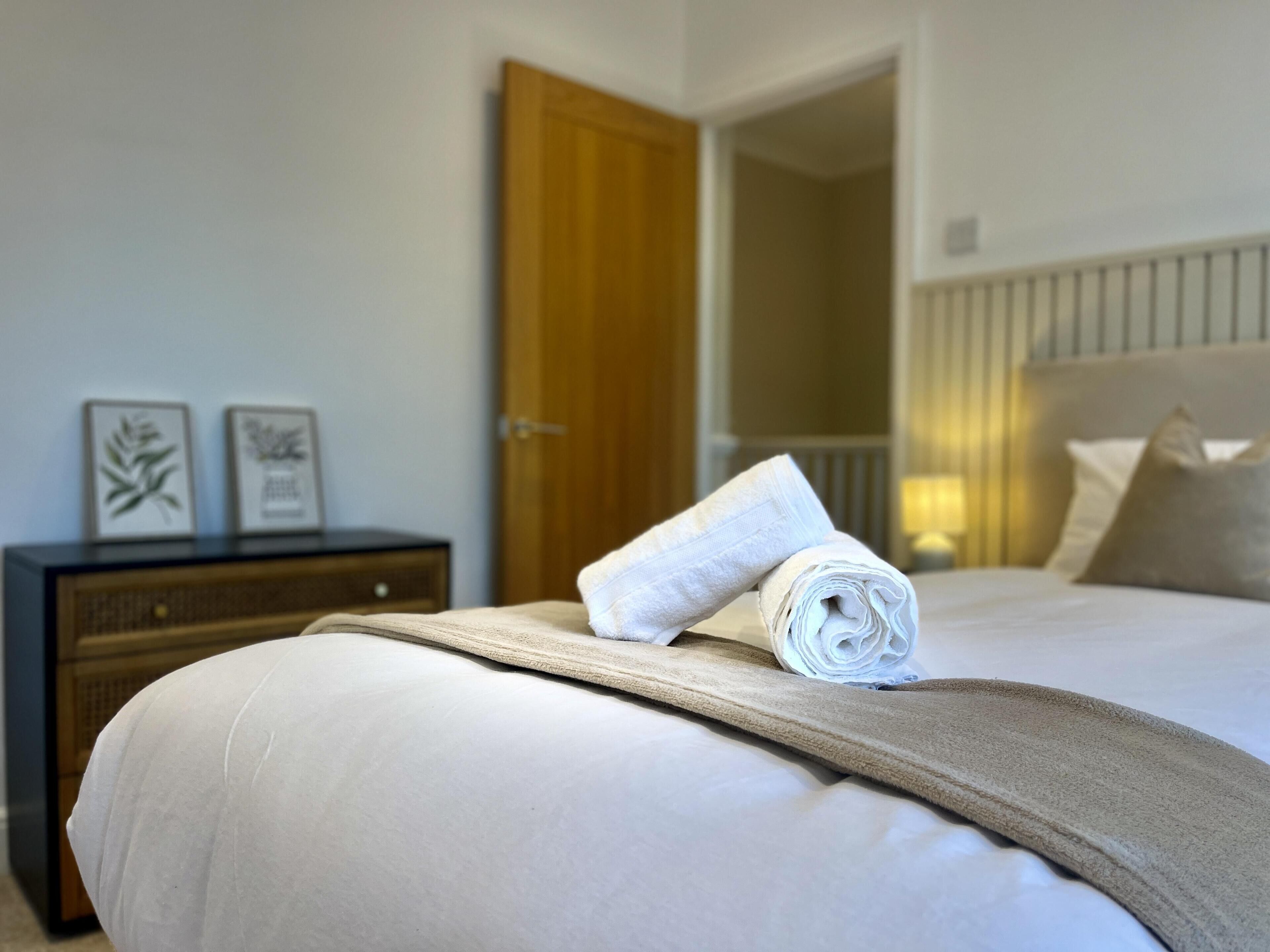 Een netjes opgemaakt bed met een opgerolde witte handdoek, een nachtkastje van hout met twee ingelijste foto’s, en een houten deur op de achtergrond.
