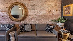 Ein modernes Wohnzimmer mit Backsteinwand, einem runden Spiegel, einem Ledersofa mit gemusterten Kissen, einem Beistelltisch mit Pflanze und einer dekorativen Lampe.