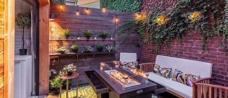 A cozy outdoor seating area with a fire feature, wooden shelves with plants, and a brick wall with ivy.