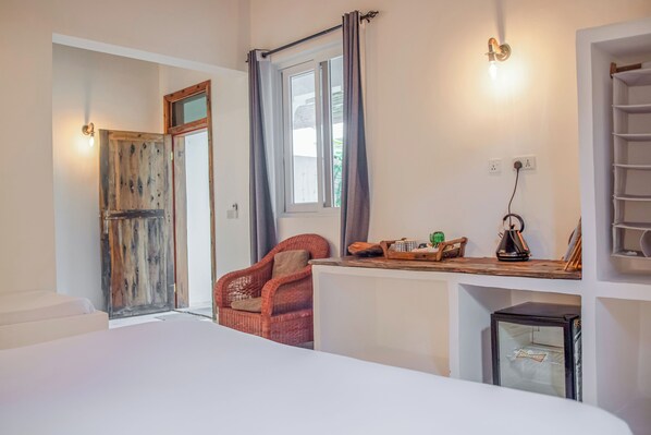 A hotel room with a bed, a wicker chair, a small table with a kettle, and a shelf with a kettle and a glass. - Cactus Lodge Zanzibar (Nungwi)