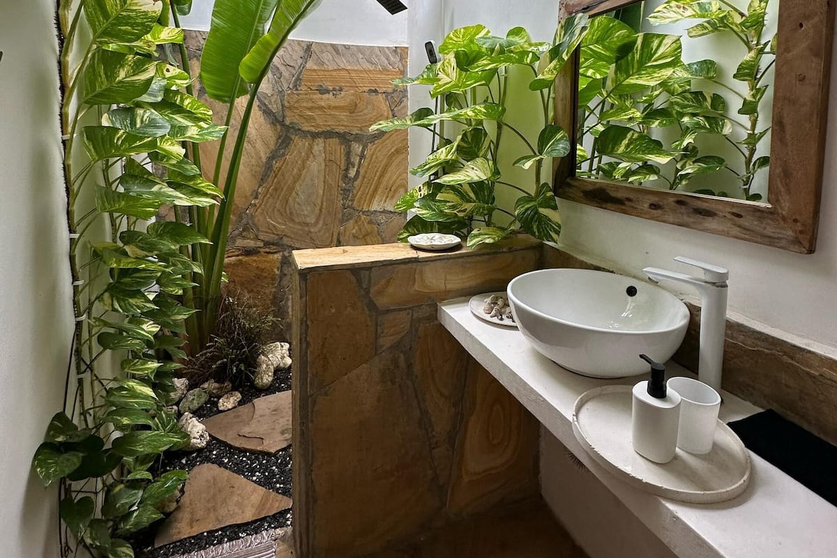 Baño con lavamanos de piedra, tocador de madera y un espejo grande. Varias plantas rodean el área del lavamanos.