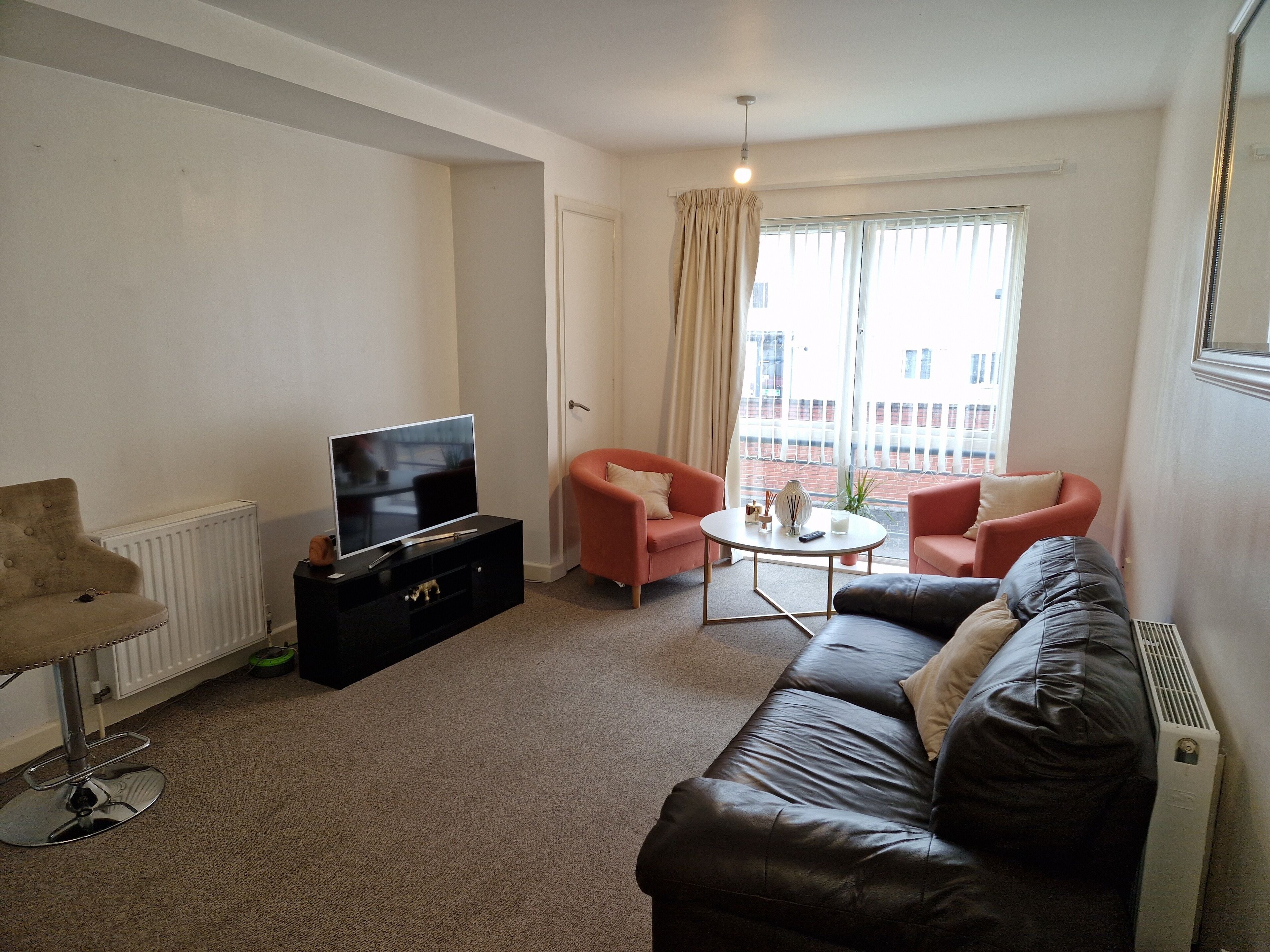 A living room with a black leather sofa, two pink armchairs, a coffee table, a TV on a black stand, and a radiator.