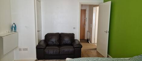 Quarto com sofá de couro marrom, cama estampada, parede verde e chão de madeira.