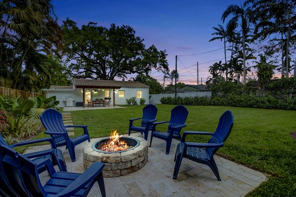 A backyard with a fire pit surrounded by blue chairs, a house in the background, and a well-maintained lawn.