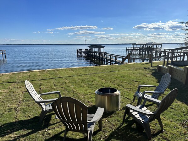 Una terraza con césped, sillas Adirondack y un pozo de fuego, con vistas a un lago tranquilo con muelles y un muelle.