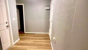 En hall med trägolv, vita väggar och en taklampa. En dörr på vänster sida leder in till ett mörkt rum.