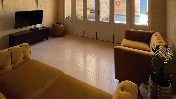 A spacious living room with a television, sofas, and a large arched window. - House with pool for 10 persons, central yet quiet area (Nadur)