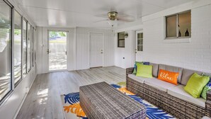 Una habitación amplia con un gran ventanal corredero que da a un patio, un ventilador de techo, un sofá de mimbre con cojines y una alfombra estampada.