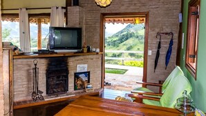 A cozy room with a fireplace, a television, and a view of a green landscape. - Pousada Dona Manoela (Gonçalves)