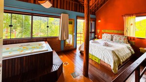 A room with a wooden ceiling, a large window with a view of greenery, a bed with floral bedding, and a bathtub. - Pousada Dona Manoela (Gonçalves)