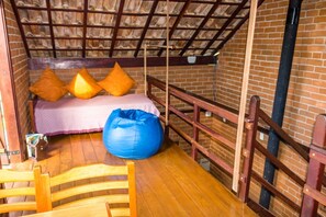 A cozy room with a wooden floor, a bed with orange pillows, a blue bean bag, and a brick wall. - Pousada Dona Manoela (Gonçalves)