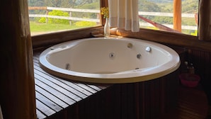 A wooden cabin with a large bathtub under a window, offering a view of mountains and greenery. - Pousada Dona Manoela (Gonçalves)