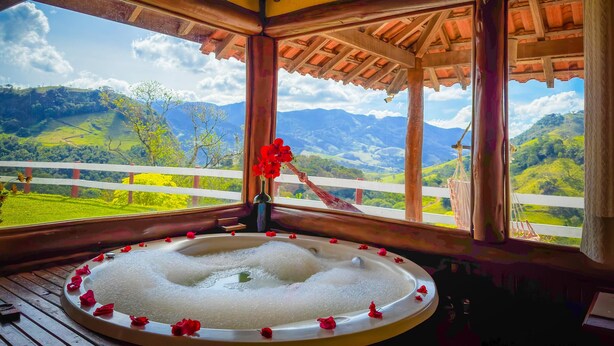 A hot tub with rose petals, a wooden deck, and a view of green hills and mountains. - Pousada Dona Manoela (Gonçalves)