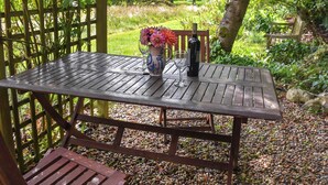 Una mesa de madera con un florero y dos botellas, sobre un suelo de grava, con una valla de madera y follaje al fondo.