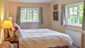 Un dormitorio con una cama grande, una mesita de noche con lámpara y jarrón, dos ventanas con cortinas y un radiador.