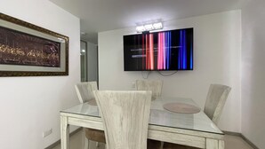 A dining area with a glass table and four chairs, a framed picture on the wall, and a television mounted above the table displaying colorful vertical lines. - Apartamento de 2 Habitaciones en Cali (Cali)