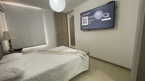 A modern bedroom with a bed, a bedside lamp, a window with blinds, a wardrobe, and a TV mounted on the wall displaying a QR code for app ID confirmation. - Apartamento de 2 Habitaciones en Cali (Cali)