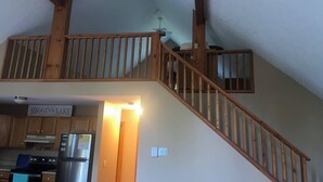 Un escalier en bois offrant une vue sur la cuisine située en contrebas, où l’on aperçoit un réfrigérateur et des placards.