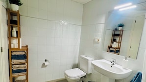 A bathroom with white tiles, a toilet, a sink, and a wooden shelf with towels.