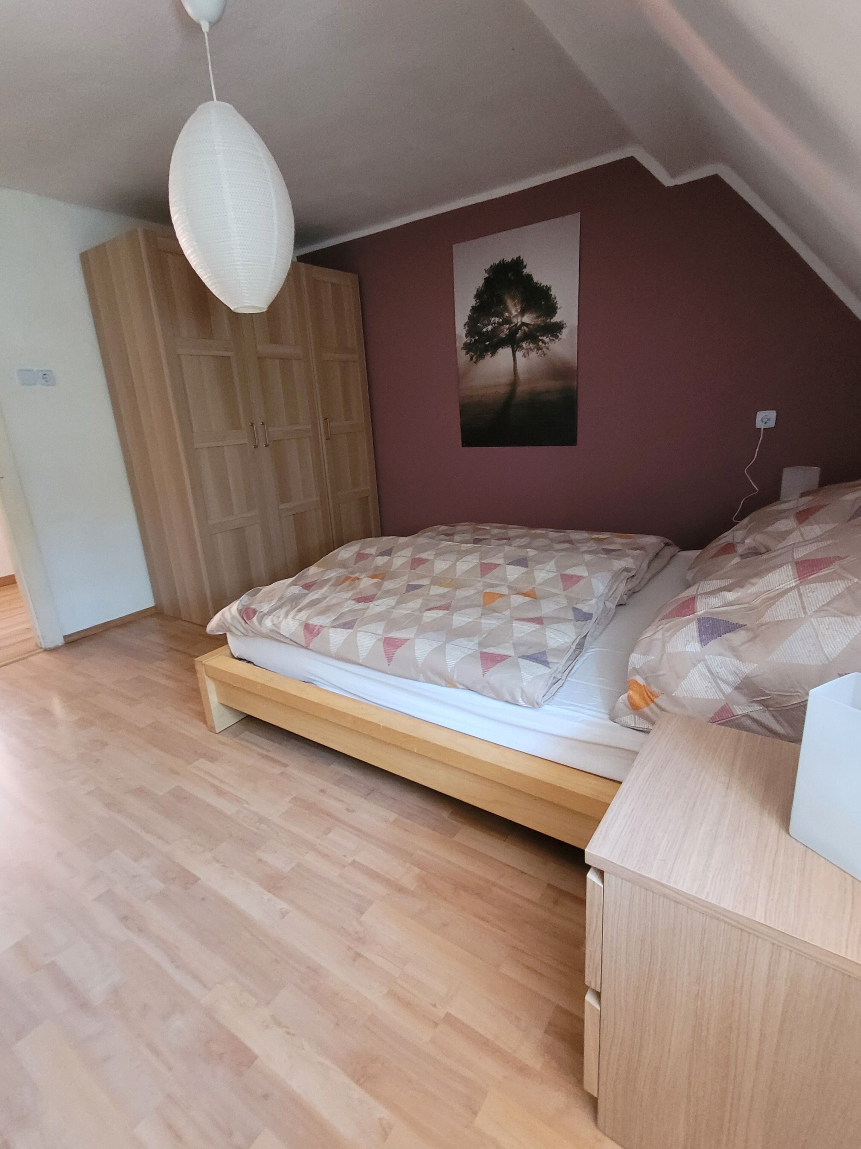 A bedroom with a wooden bed, a white pendant light, a wall-mounted picture of a tree, and a wooden wardrobe.