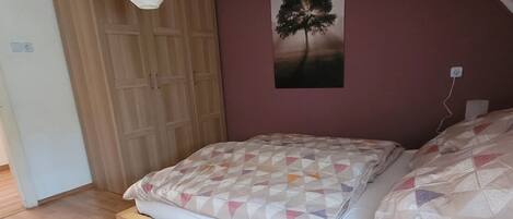 A bedroom with a wooden bed, a white pendant light, a wall-mounted picture of a tree, and a wooden wardrobe.