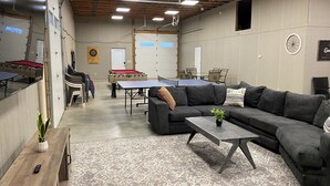 Un espacio interior amplio con mesa de ping pong, mesa de fútbolín y un sofá. Hay sillas y un dardo en las paredes.