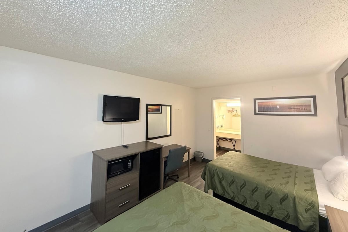 A hotel room with two single beds, a desk with a chair, a television mounted on the wall, and a small kitchenette area.