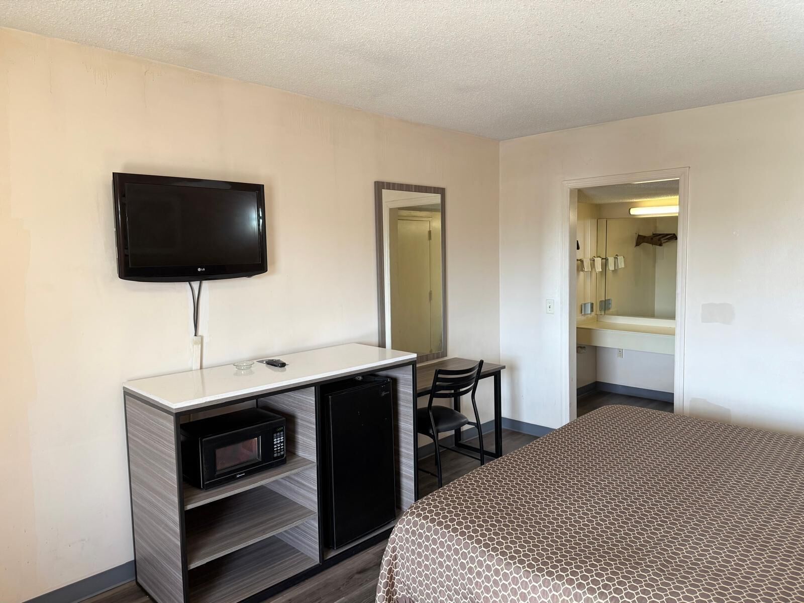 Habitación de hotel con cama, escritorio con microondas, refrigerador, televisor, espejo y baño visible a través de una puerta abierta.