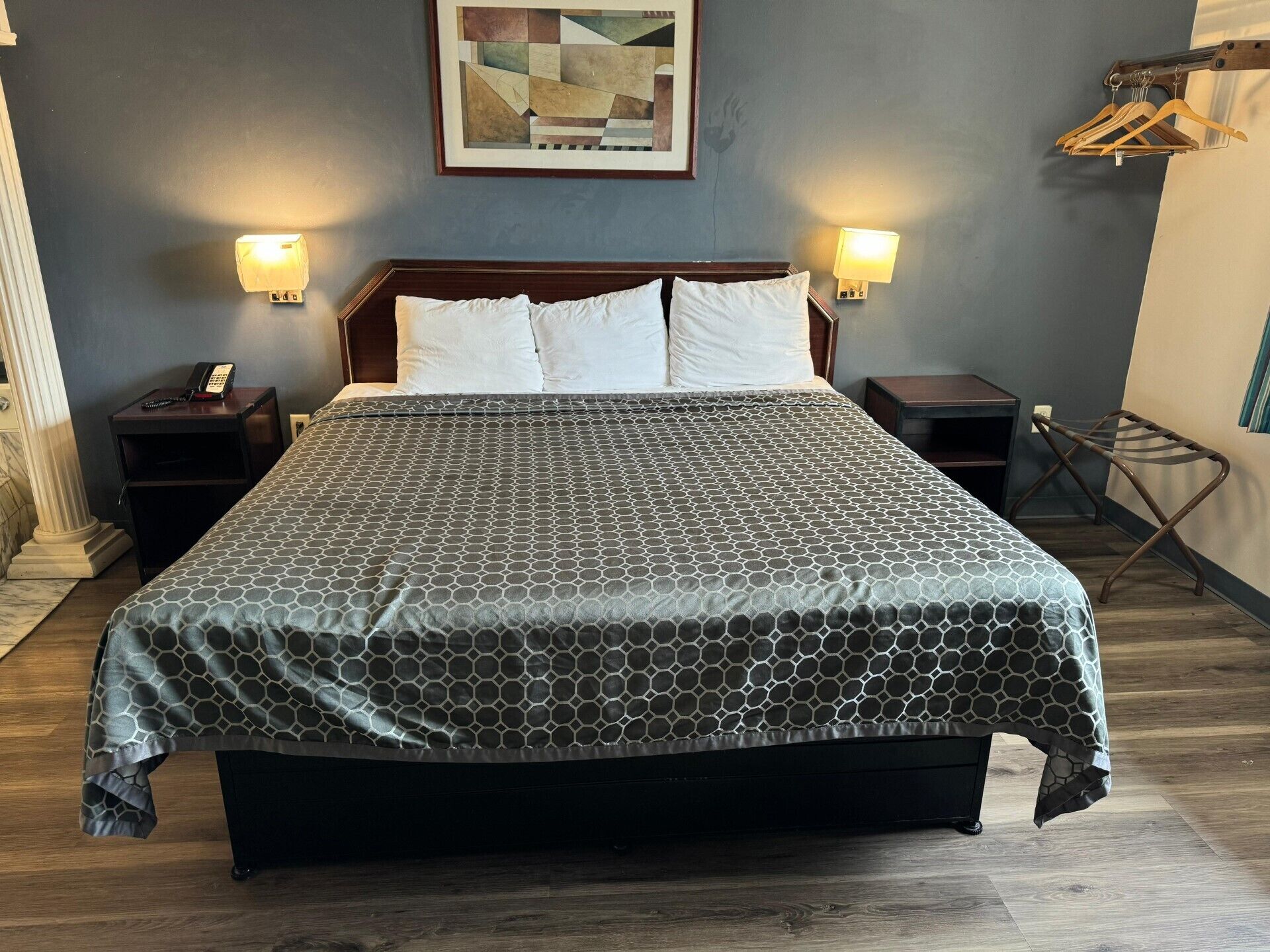 Un letto rifatto con cura, con copriletto a motivi geometrici, due comodini con lampade, un quadro astratto incorniciato a parete e un appendiabiti in legno.