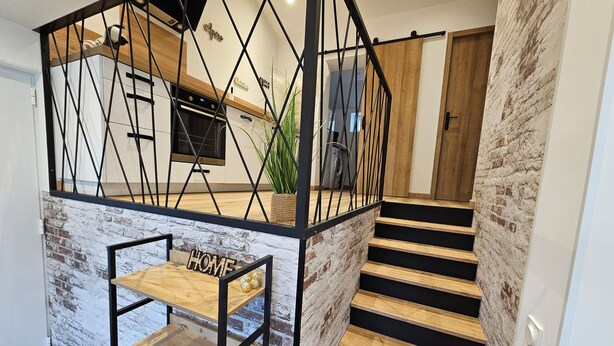 Una escalera moderna con detalles de madera y barandillas negras, que conduce a una zona de cocina con un ventanal de marco negro y una planta.