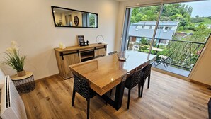 Un comedor moderno con mesa y sillas de madera, una pequeña cocina y un amplio ventanal con vistas a un barrio residencial.