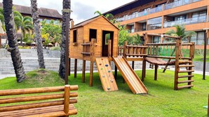 Estructura de juego de madera con tobogán y pared de escalar, rodeada de césped y palmeras.