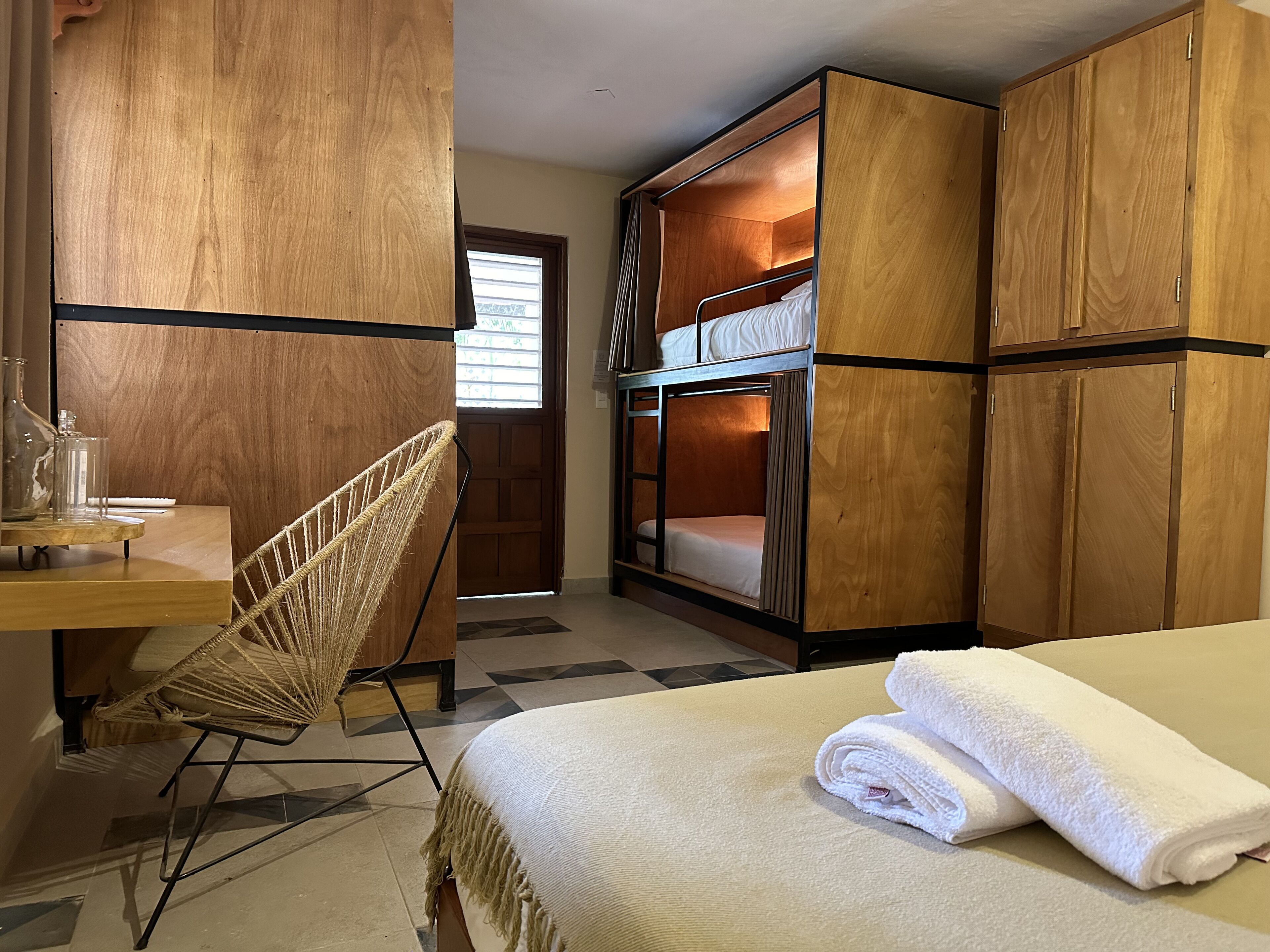 A compact room with a bunk bed, wooden wardrobe, a desk with a chair, and a bed with rolled towels.