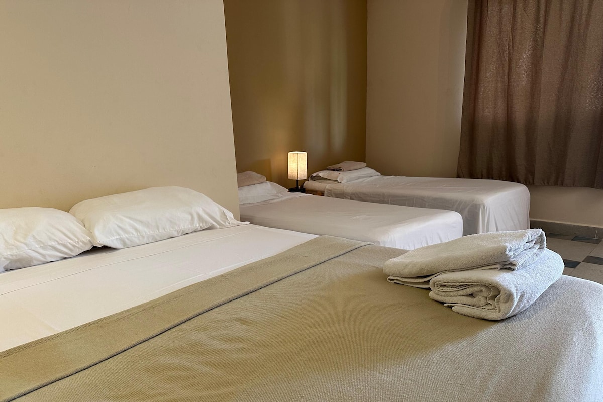 A hotel room with two beds, a large bed with a beige blanket and pillows, and a smaller bed with a white blanket and pillows. There is a lamp on a bedside table and a folded towel on the large bed.