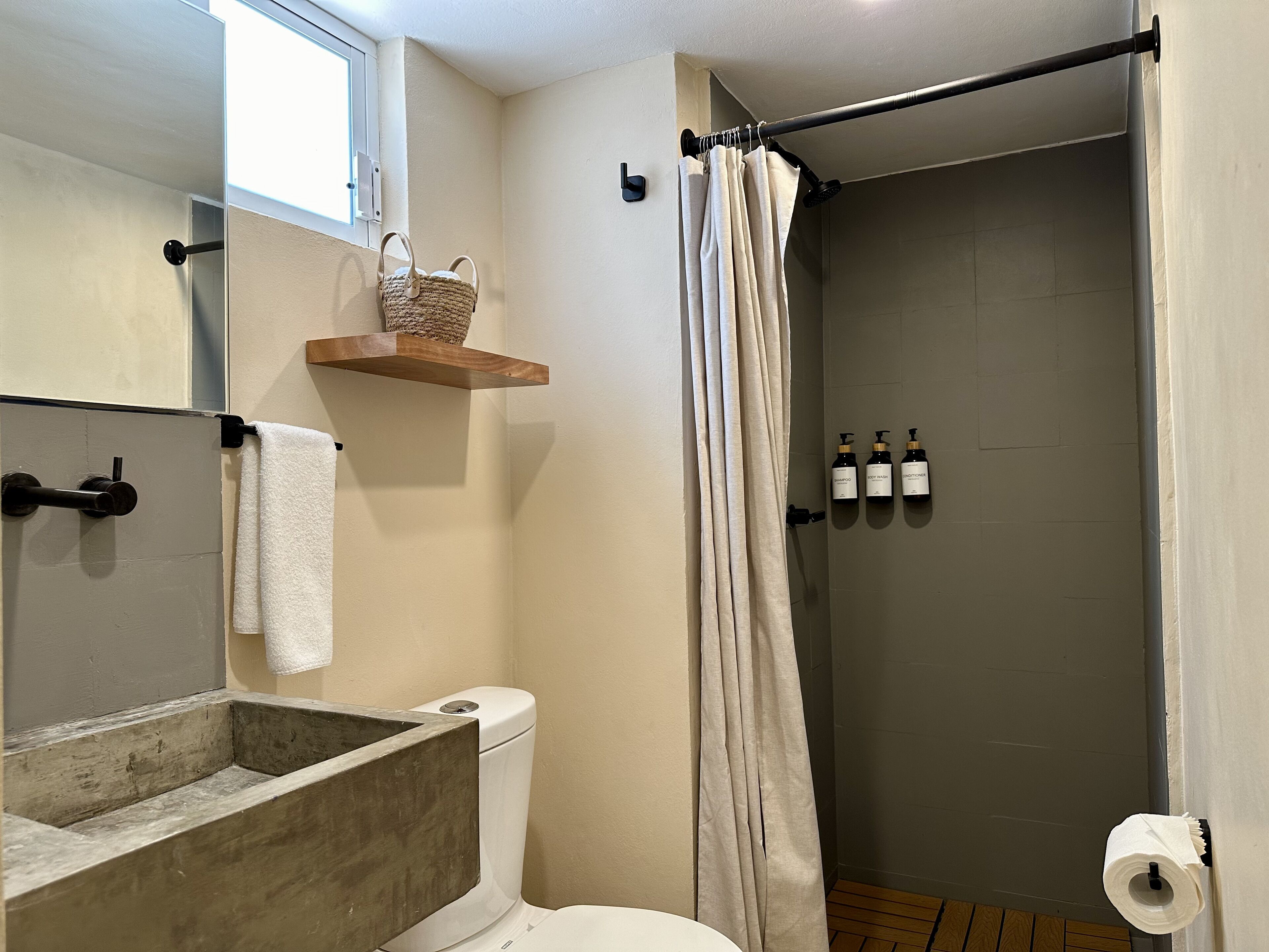 A bathroom with a toilet, a wooden shelf with a basket, a towel, a shower curtain, and three bottles of shampoo.