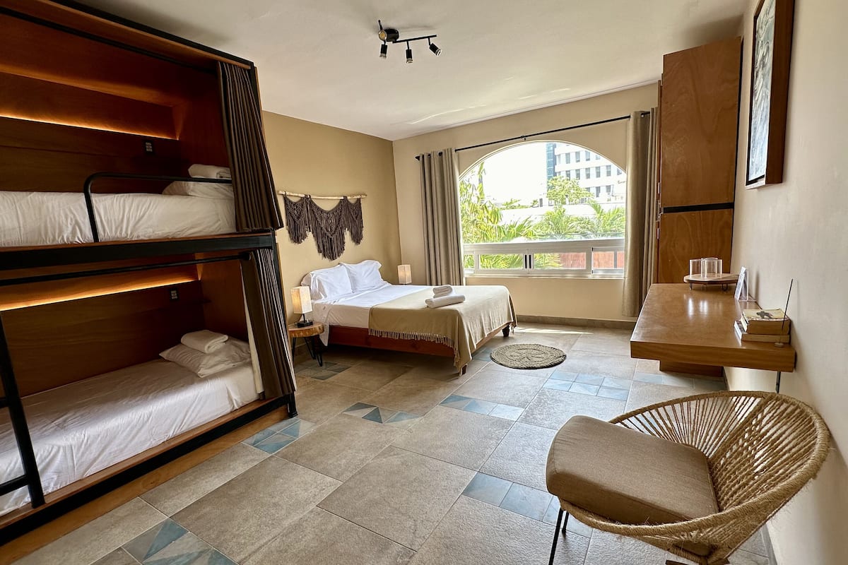 A hotel room with bunk beds, a single bed, a round woven chair, a small table, and a view of greenery and buildings through the window.
