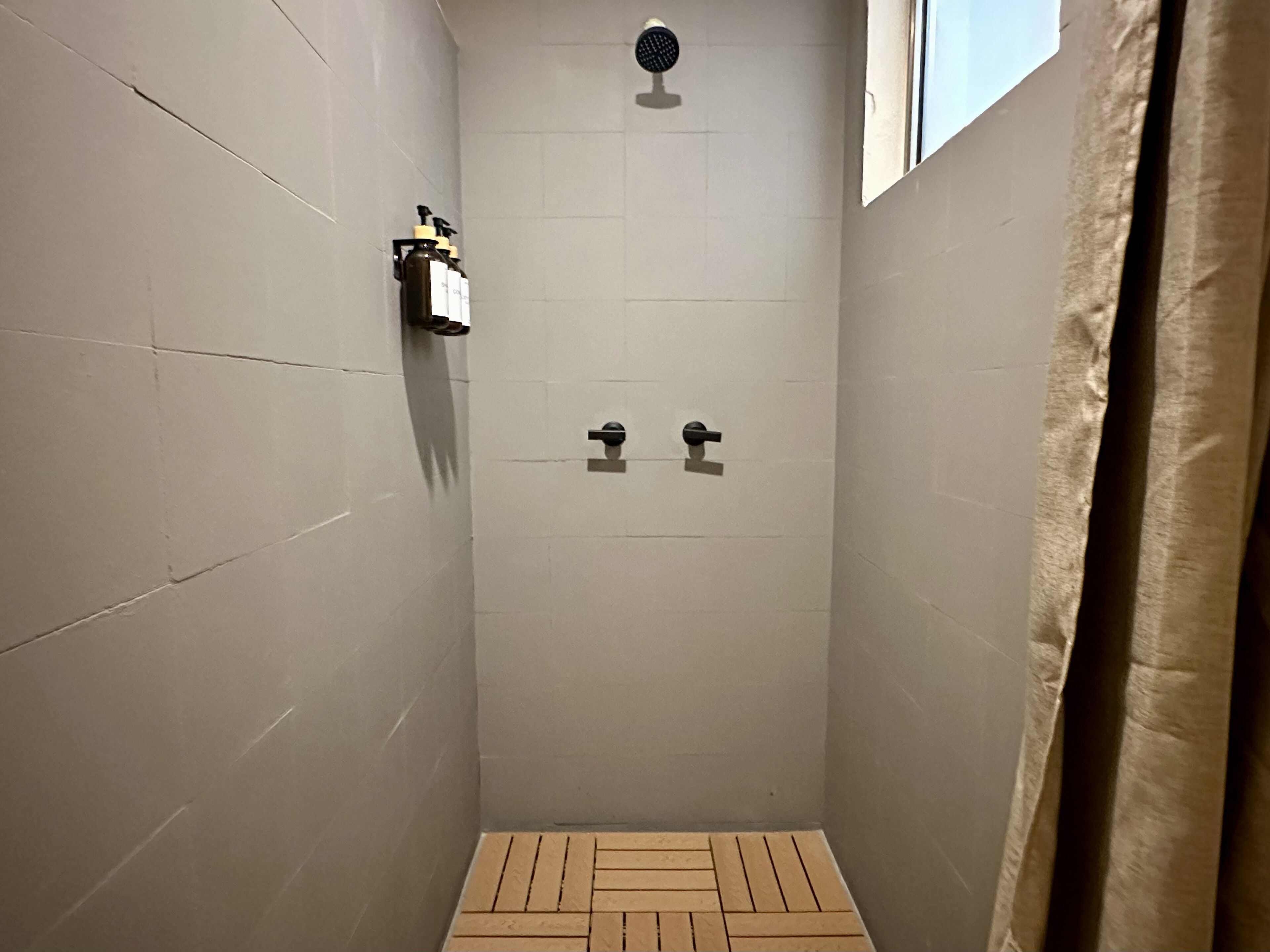A small, tiled shower room with a single showerhead, a towel dispenser, and a window.