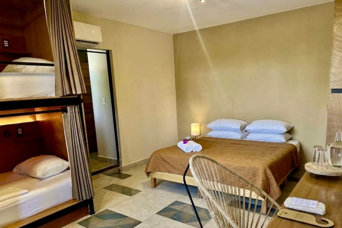 A hotel room with bunk beds, a single bed, a chair, a table with a bottle and a glass, and a lamp.