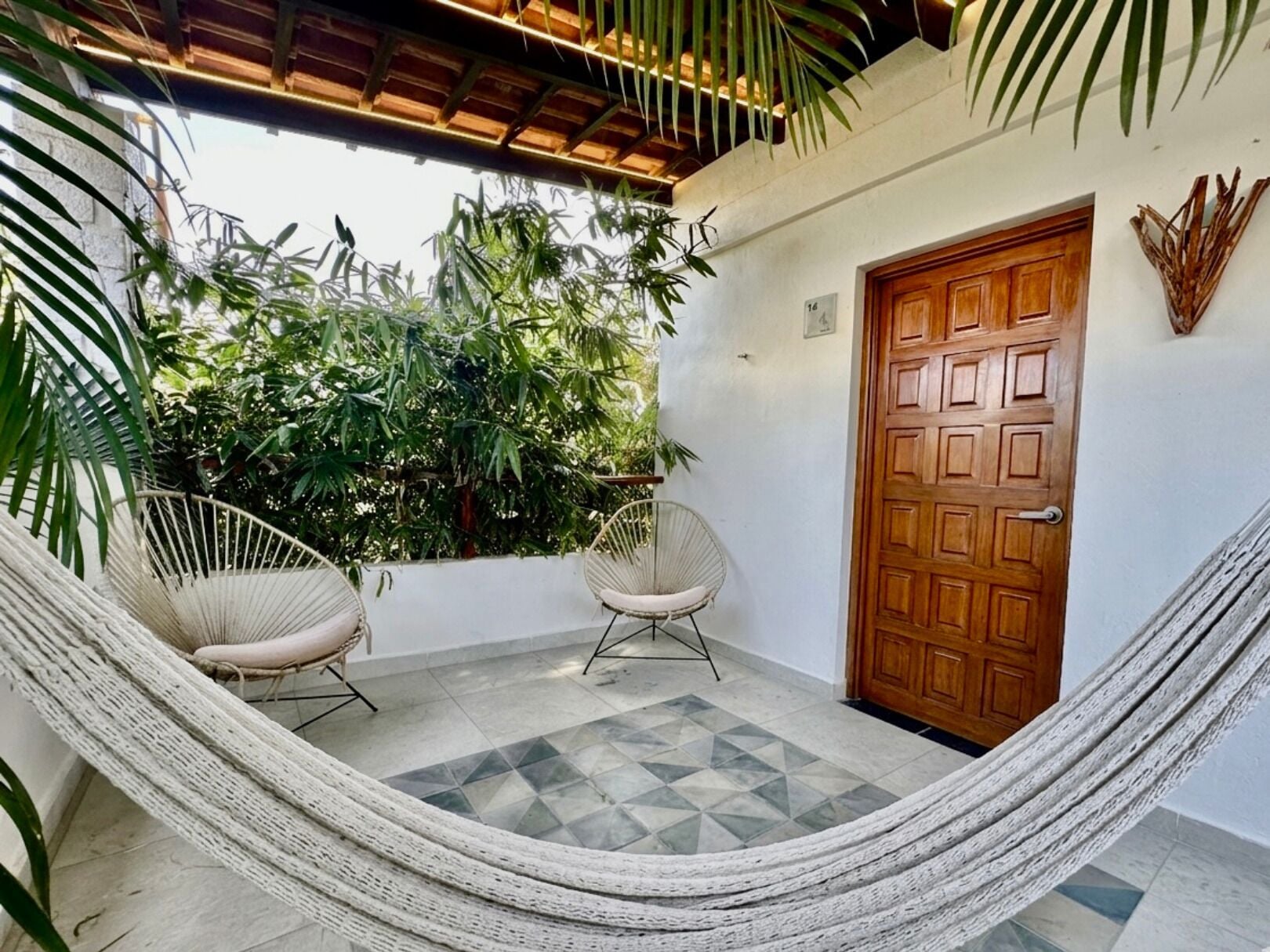 A hammock, two wicker chairs, a wooden door, and a tiled patio with greenery in the background.