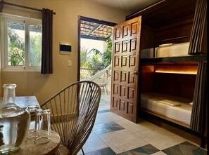 A room with a bed, a chair, a table with glasses, and a door leading to a balcony with plants.
