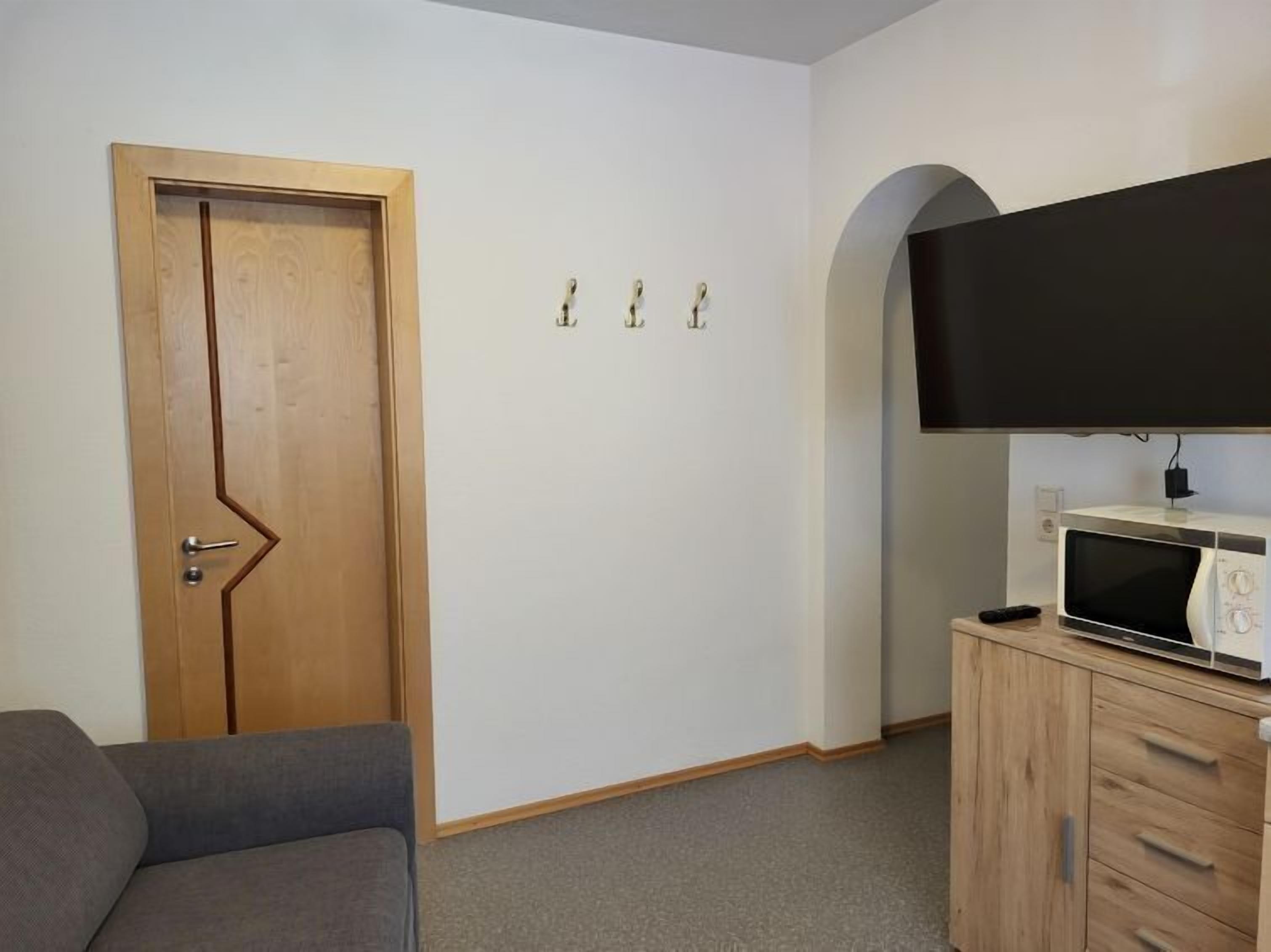 A room with a wooden door, a grey sofa, a microwave on a wooden cabinet, and a flat-screen TV mounted on the wall.