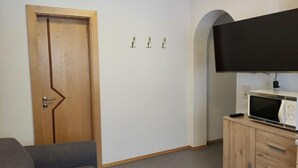 A room with a wooden door, a grey sofa, a microwave on a wooden cabinet, and a flat-screen TV mounted on the wall. - Christians Hamat by Interhome (Ischgl)