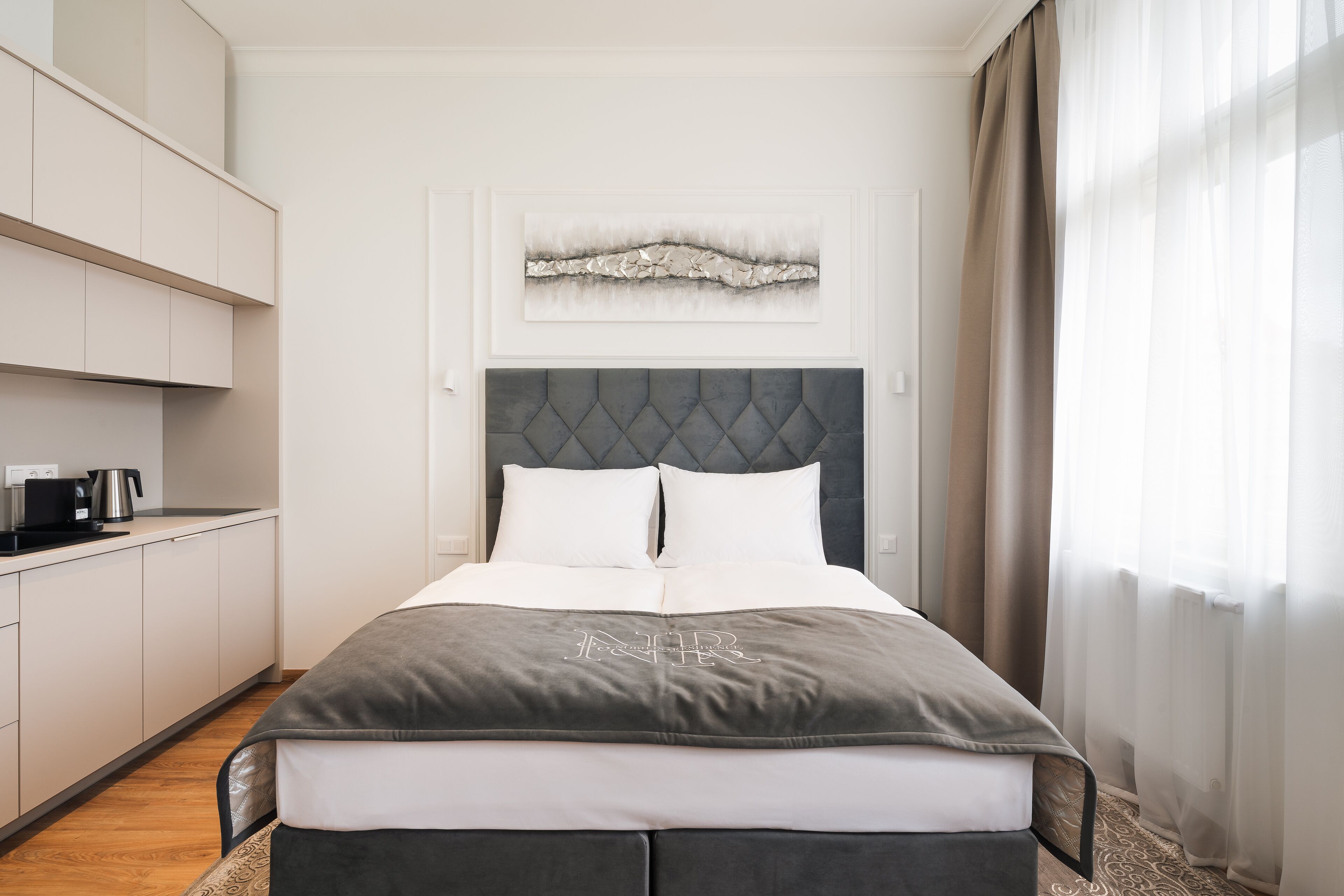 A modern hotel room with a bed, a kitchenette, and a painting on the wall.