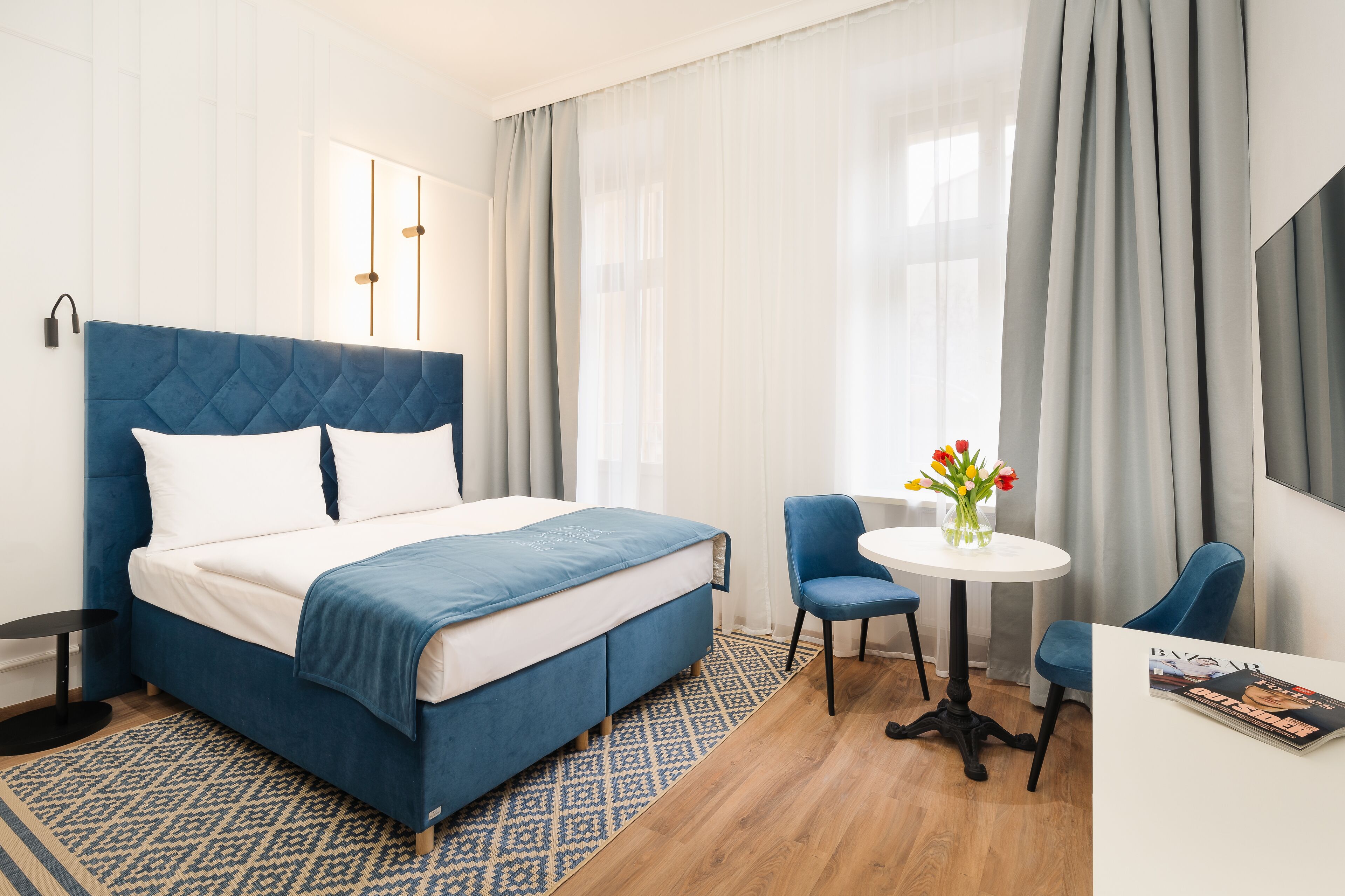 A modern hotel room with a large bed, a small round table with chairs, and a large window with curtains.