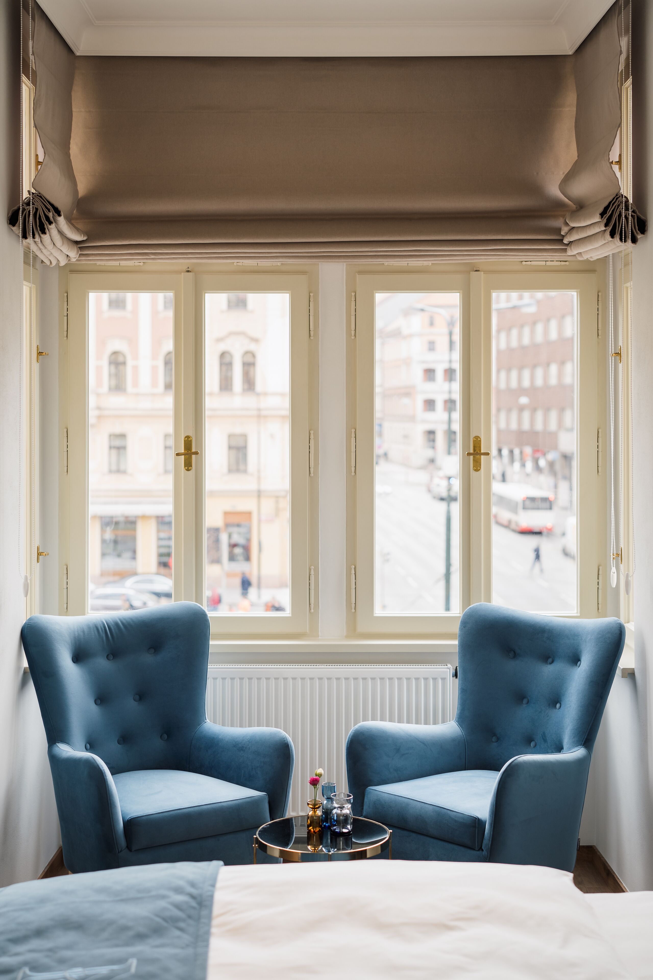 Une pièce avec une grande fenêtre, deux fauteuils bleus, une petite table ronde avec un plateau, et un lit recouvert d’une couette blanche.