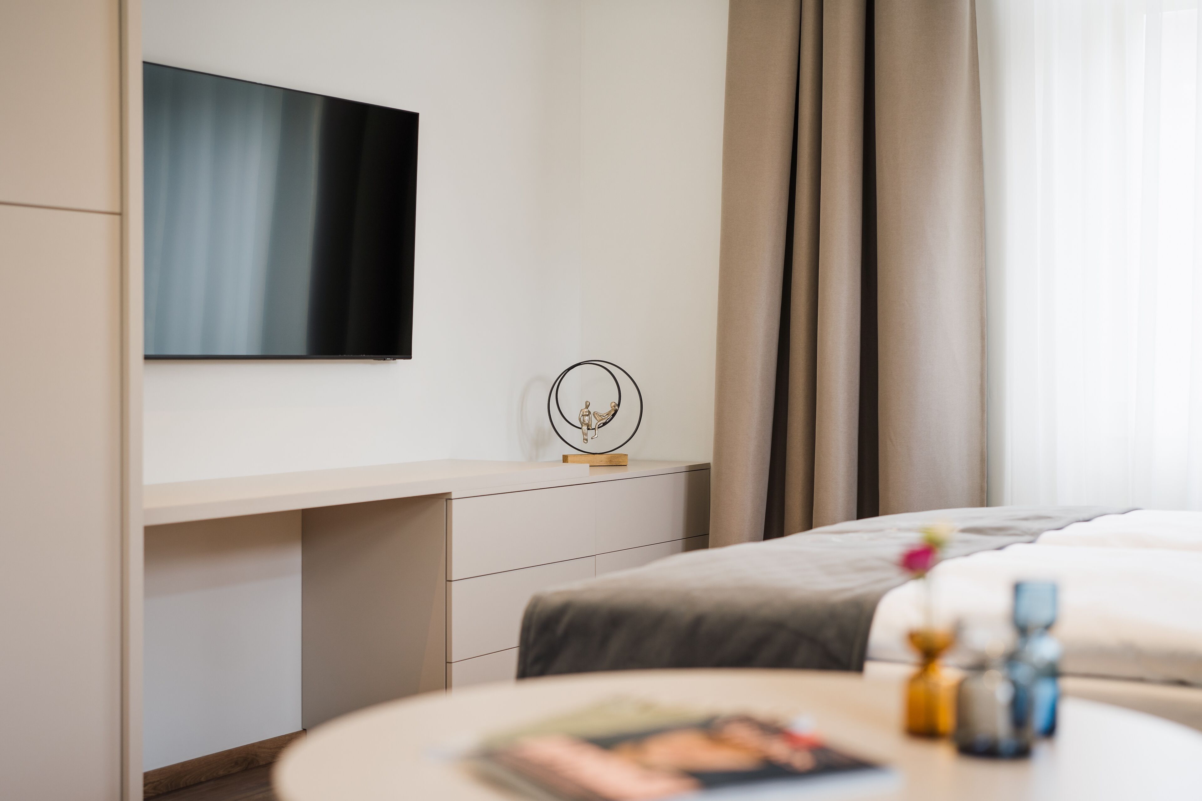 Et moderne hotelværelse med et fladskærms-tv på væggen, et natbord med en dekorativ genstand, en seng med et gråt tæppe og et vindue med gardiner.